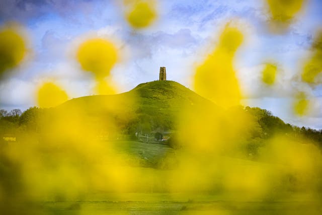 As pessoas circulam pela rua St. A torre de Michael no topo de Glastonbury Tor, vista através de colza amarela florescendo em um dia de clima misto em Glastonbury, Somerset