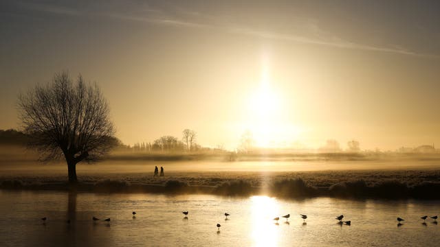People walk through frost and mist alongside a frozen lake during sunrise in Bushy Park, London