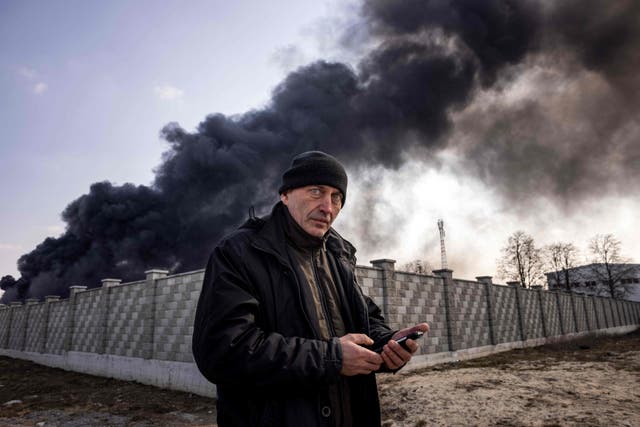 A man holds a mobile phone while smoke rises after Russian attacks hit a fuel storage facility in the city of Kalynivka