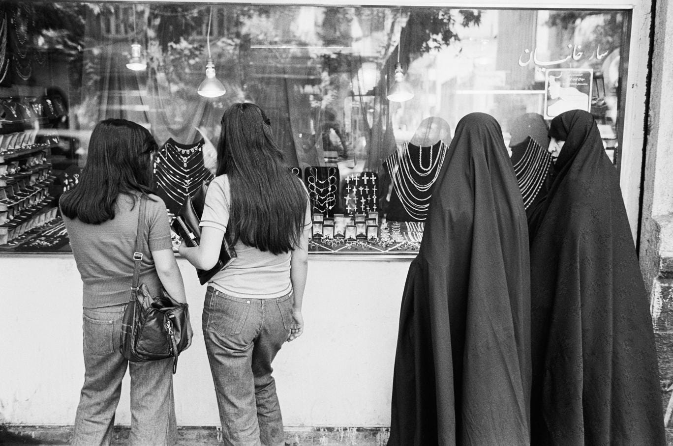 Women in traditional and western clothing stand side by side as they look at a shop window in Iran