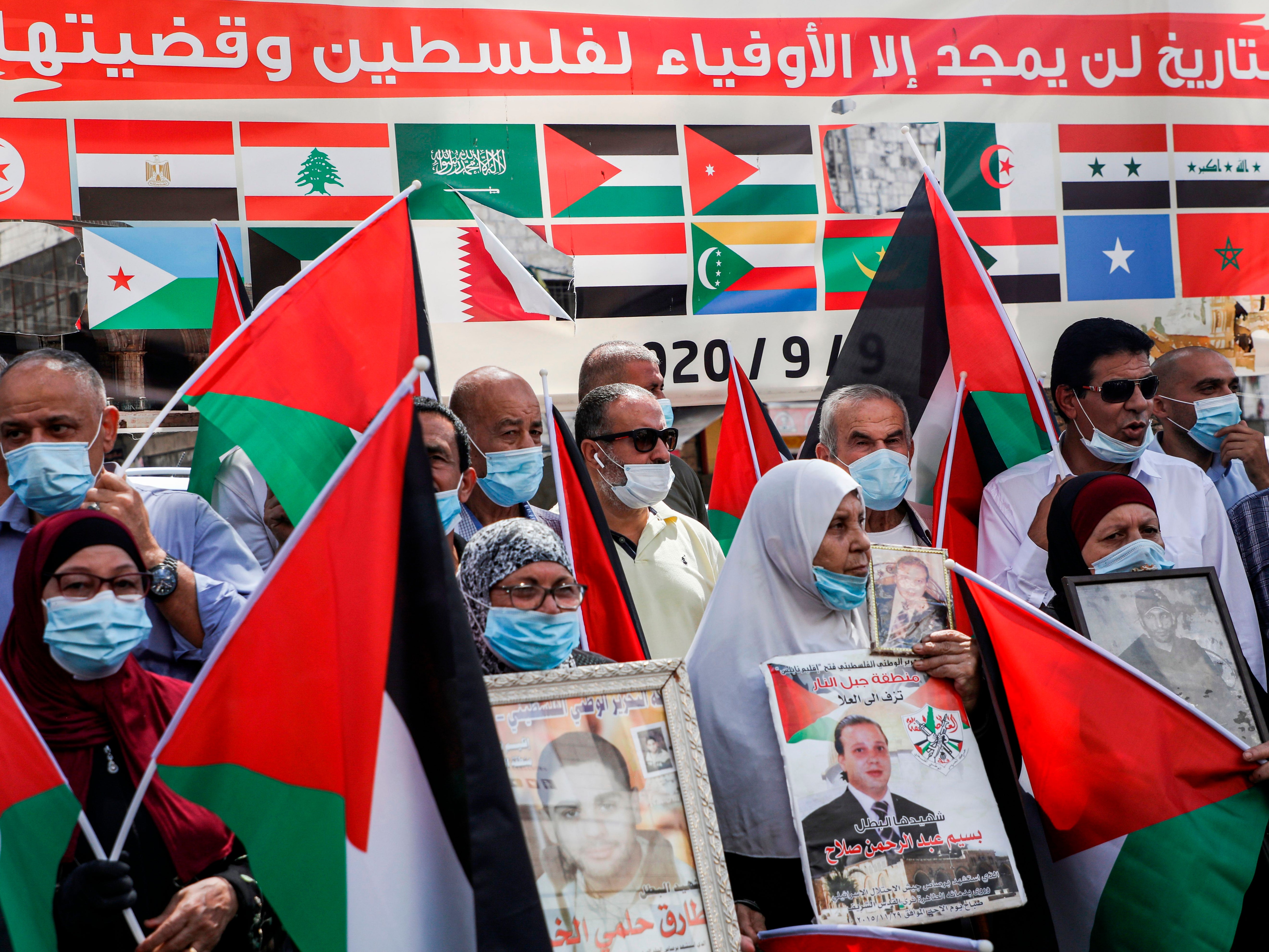 Demonstrators with Palestinian flags stand before a banner showing the flags of Arab League member states with text in Arabic above reading: 'History will glorify but those faithful to Palestine and its cause'