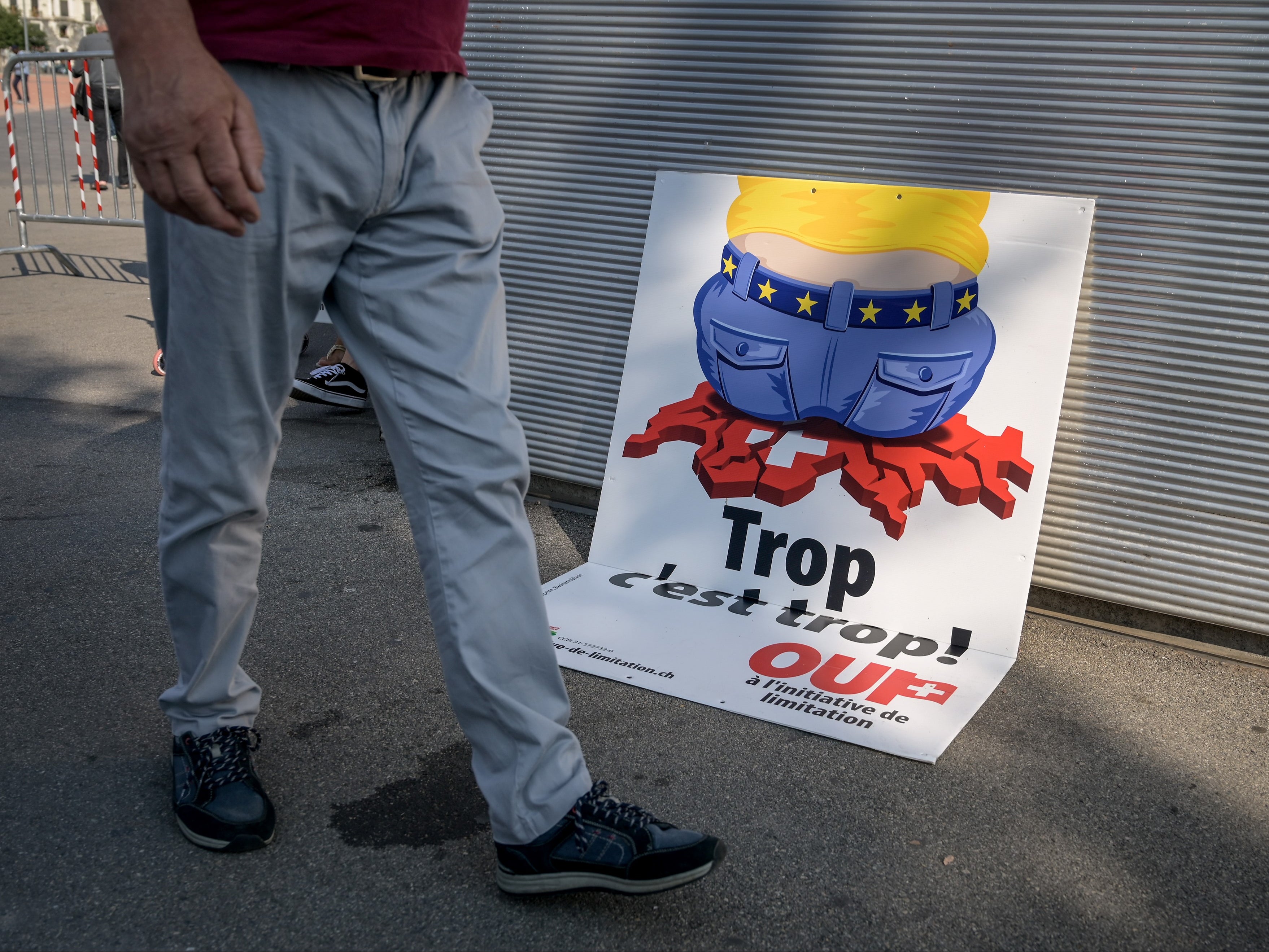 An electoral poster by right-wing Swiss Peoples Party shows a cartoon worker wearing a belt studded with EU stars, crushing the red and white map of Switzerland with his wide rear end that translates from French as 'Too much is too much!'