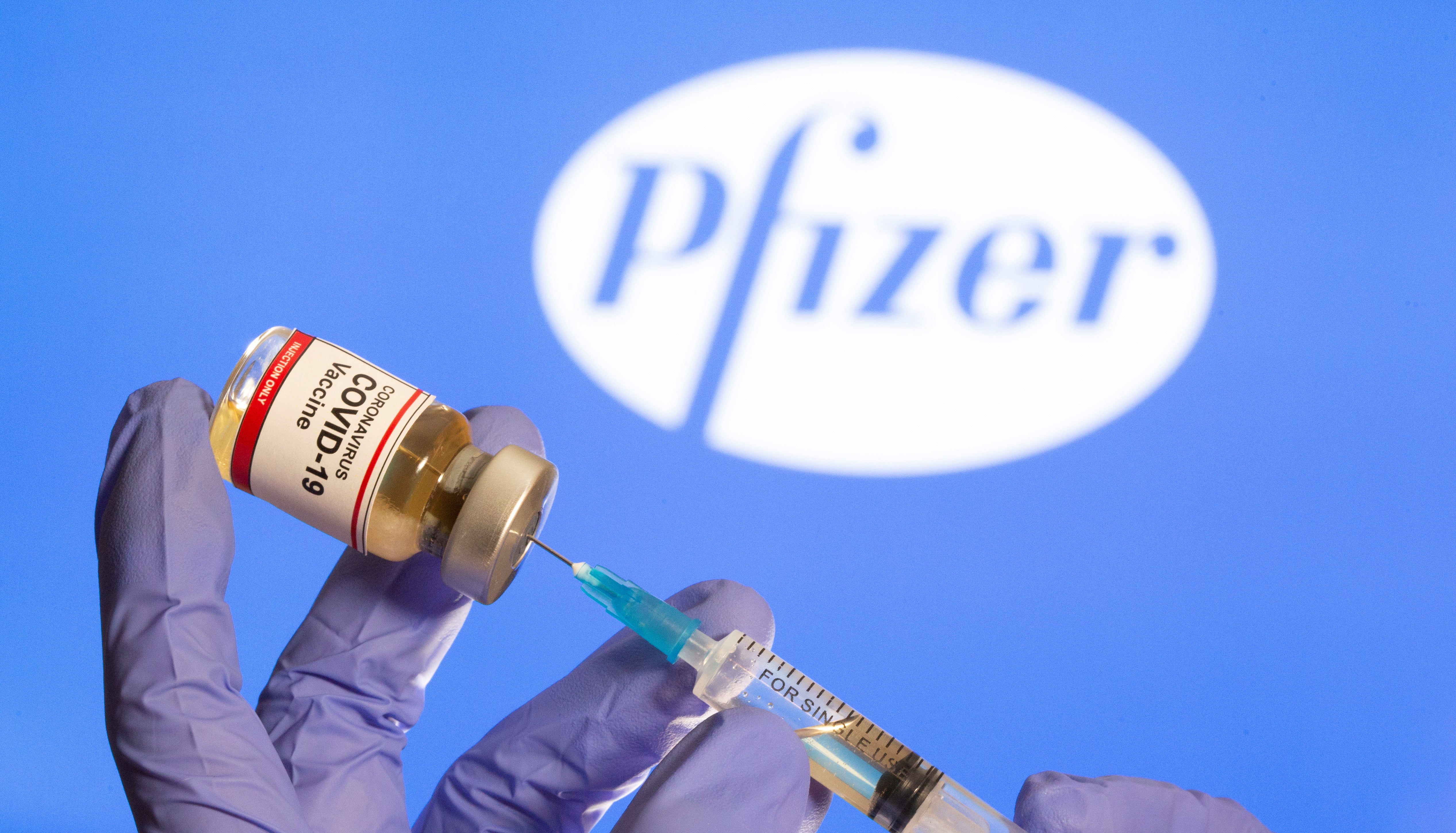 A woman holds a small bottle labeled with a “Coronavirus COVID-19 Vaccine” sticker and a medical syringe in front of displayed Pfizer logo on 30 October 2020.