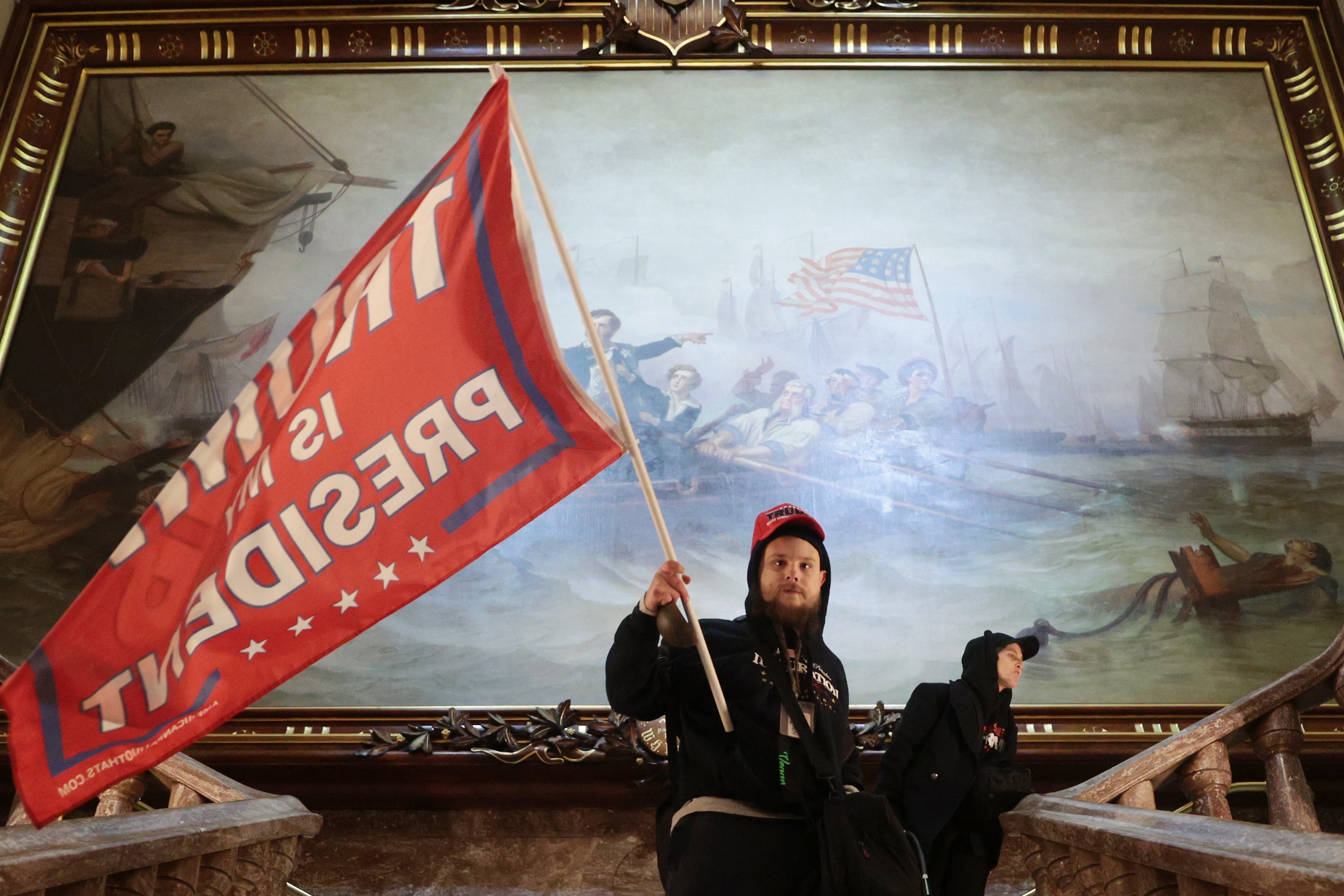 Un manifestante ondea una bandera de Trump dentro del edificio del Capitolio de los Estados Unidos frente a una pintura del siglo XIX el 6 de enero de 2021.