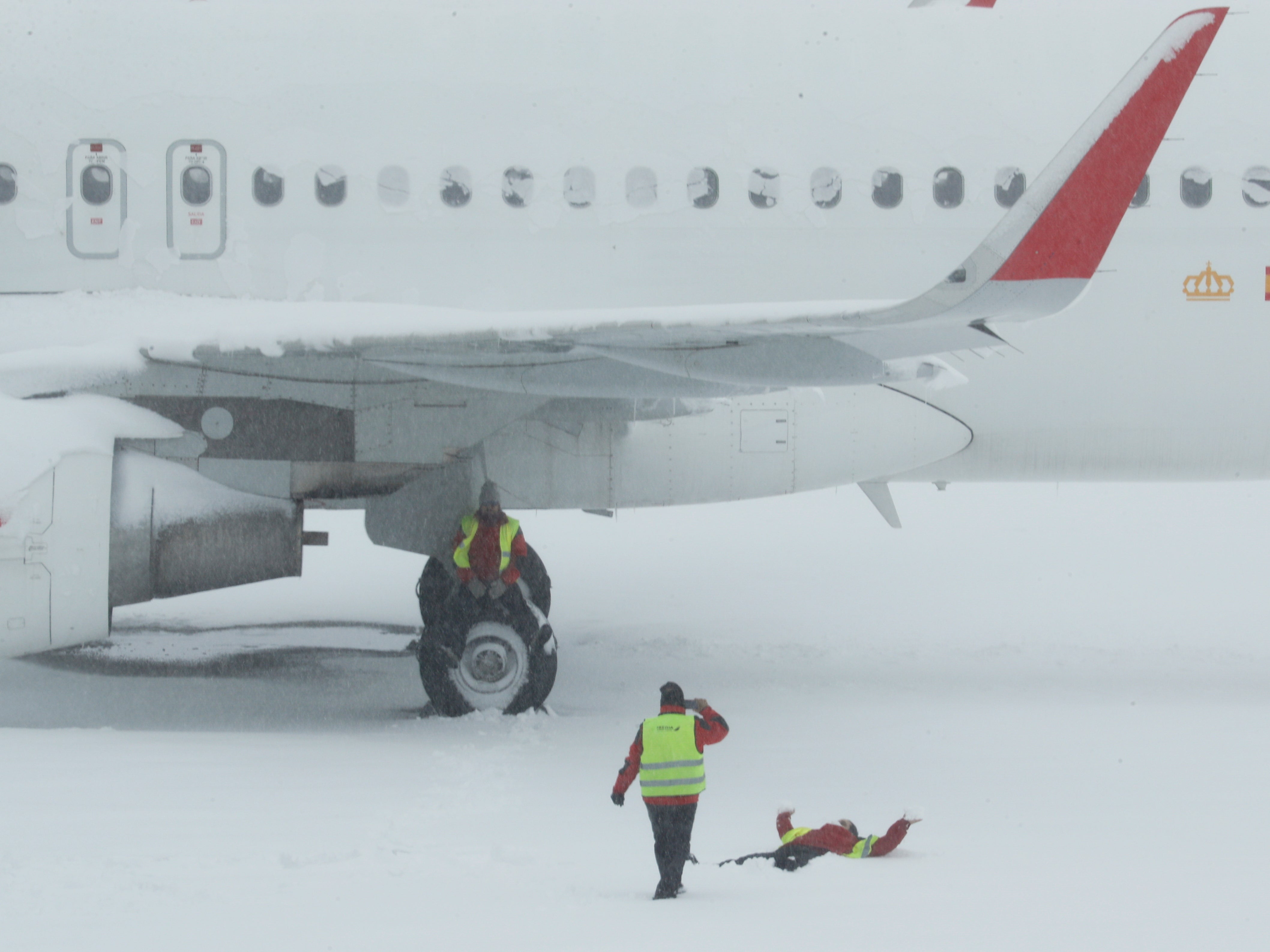 An airport worker sits on the wheel of a parked snow-covered plane as one of his colleague takes a picture of another coworker lying on the snow at Adolfo Suarez Barajas airport, which is suspending flights due to heavy snowfall in Madrid