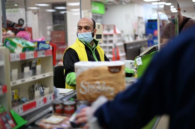 <p>A shop worker wearing a protective face covering to combat the spread of the coronavirus serves customers at an Asda supermarket in London in December&nbsp;</p>