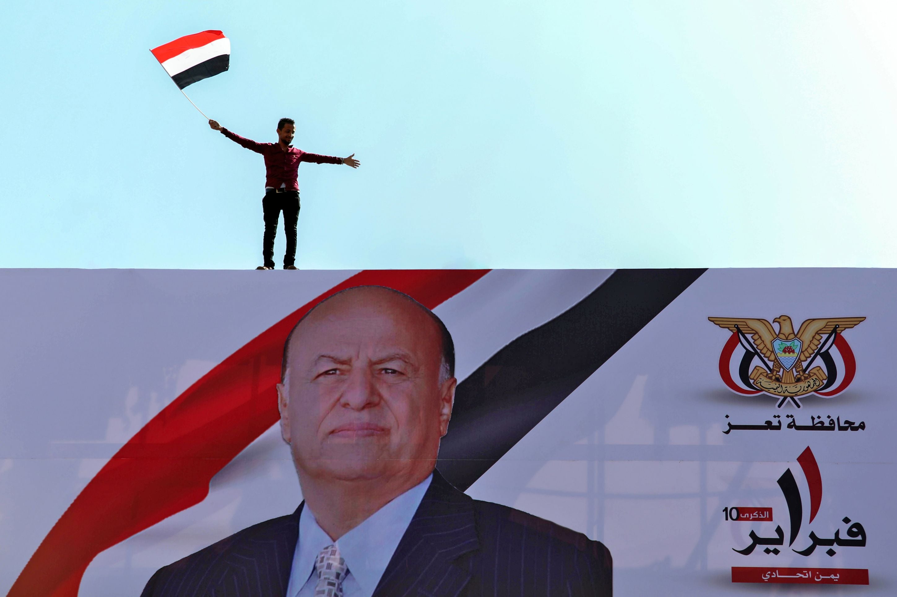 A Yemeni youth waves a national flag atop a billboard in Taez showing Yemen’s President Abedrabbo Mansour Hadi during a rally commemorating the 10th anniversary of the Arab Spring uprising