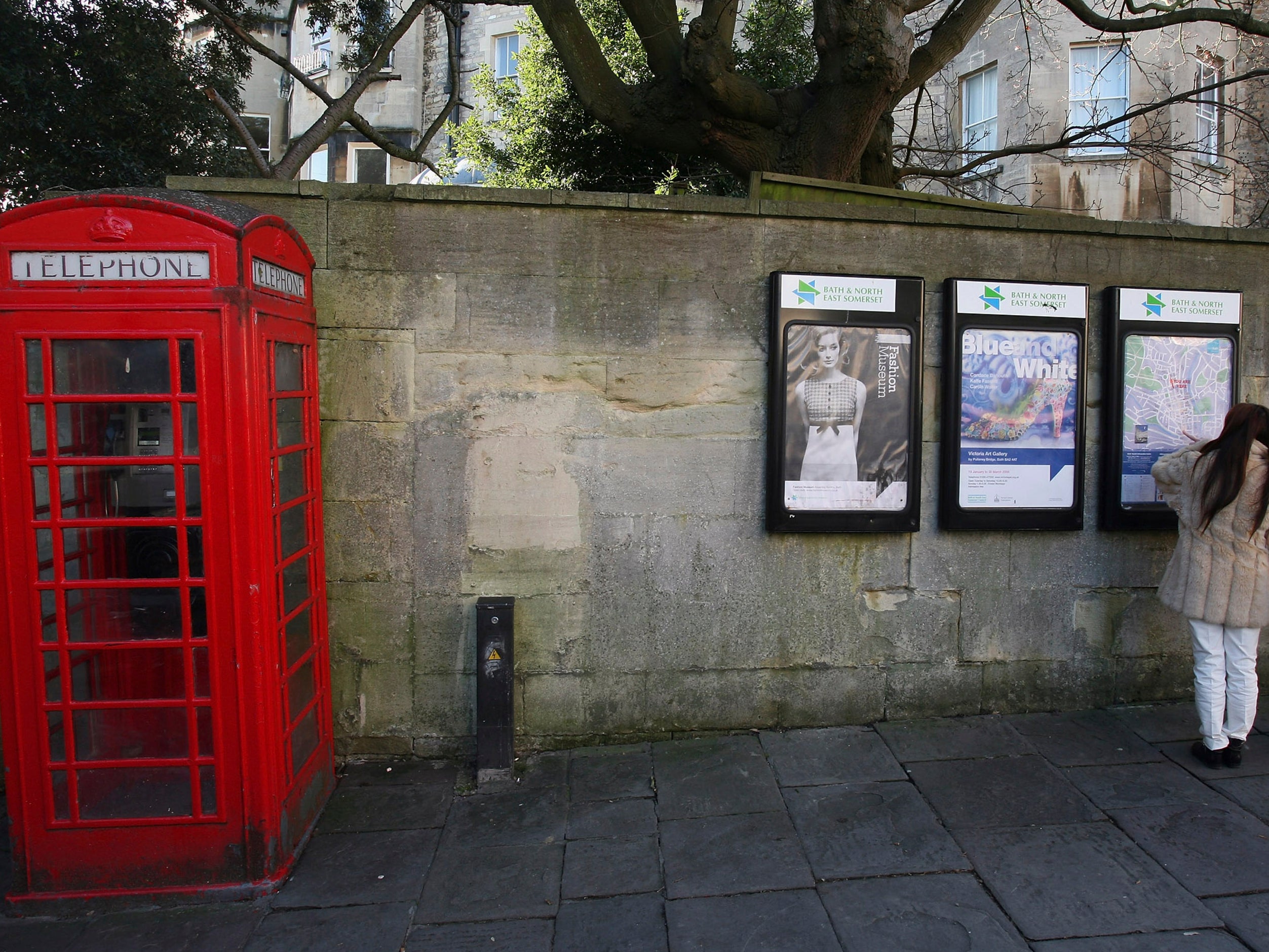 Two women look at posters on a wall beside a traditional red telephone box on a street in Bath