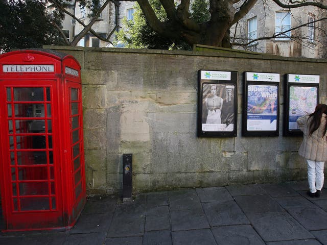 Two women look at posters on a wall beside a traditional red telephone box on a street in Bath 