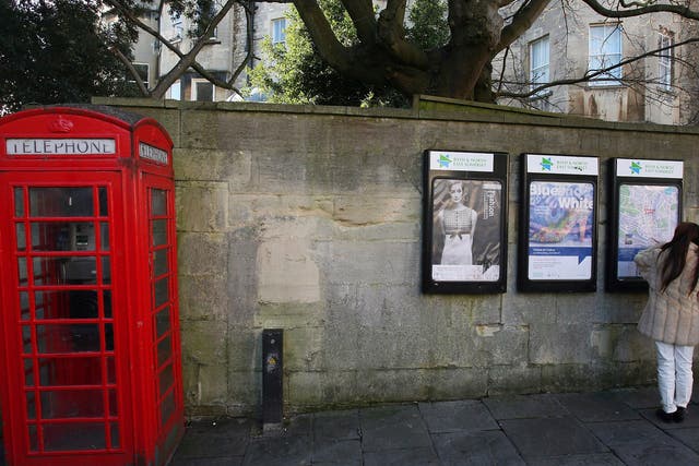 Two women look at posters on a wall beside a traditional red telephone box on a street in Bath 