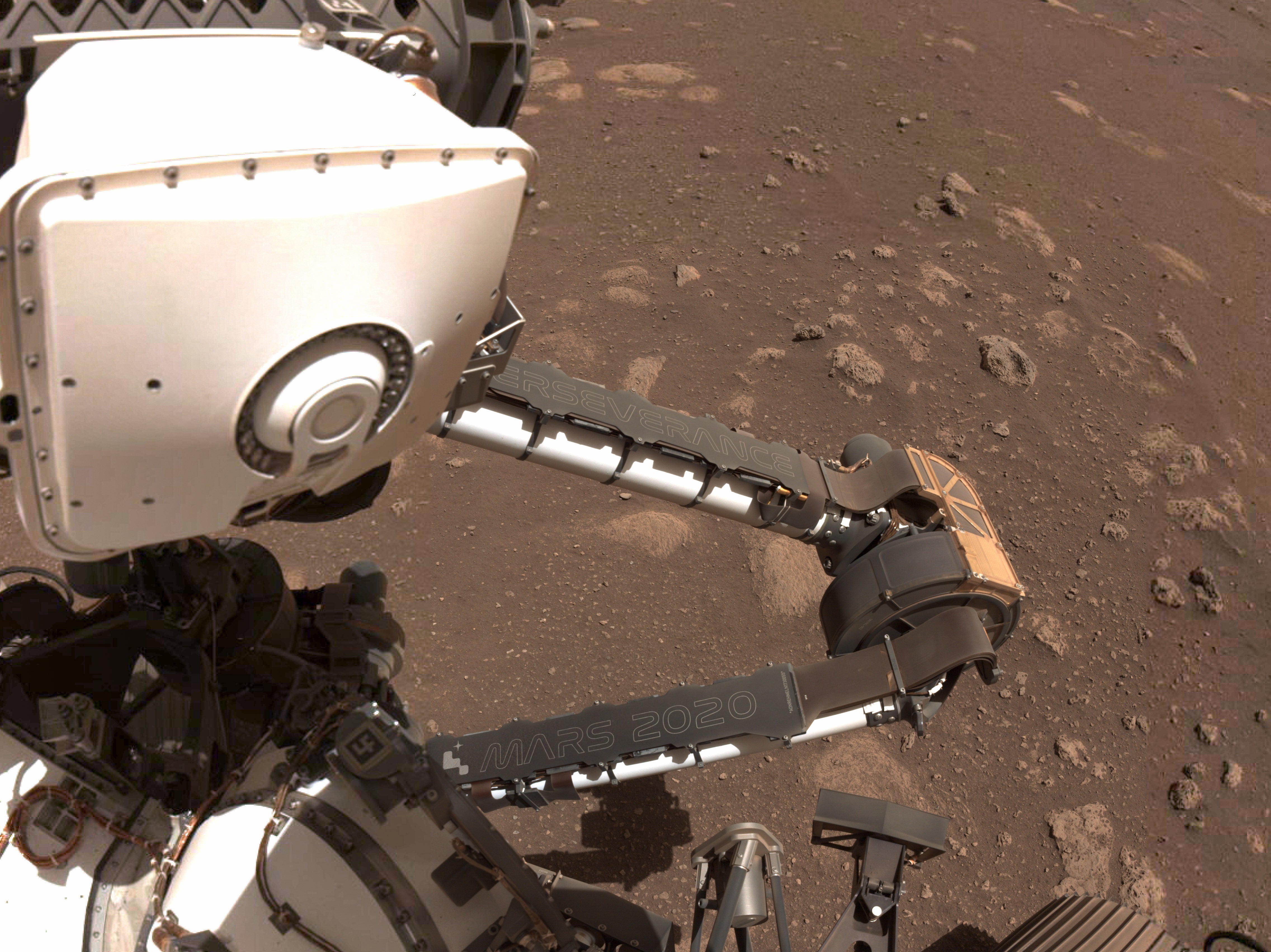 Two name plates mounted on the Mars Perseverance rover’s robotic arm. The rover’s name, “Perseverance,” is inscribed on the plate attached to its forearm, and the mission name, “Mars 2020,” is shown on its upper arm.