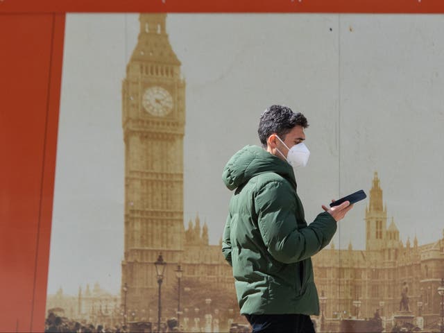 A man wearing a protective face mask looks at his smartphone as she passes a picture of the Houses of Parliament in central London