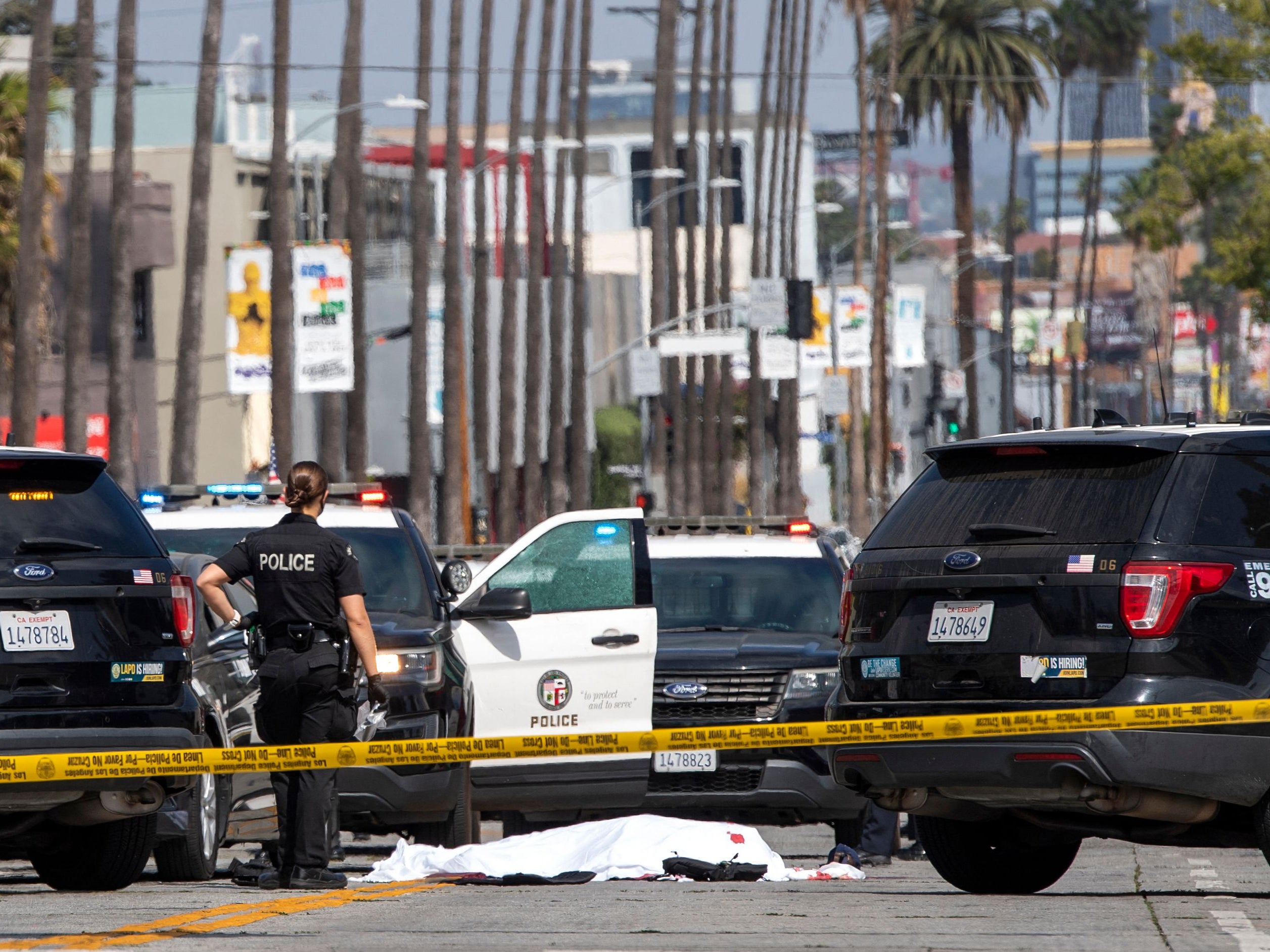 A LAPD police officer stands at the corner of Fairfax Avenue and Sunset Boulevard where a body covered in a white sheet lies on the pavement in Los Angeles on 24 April, 2021