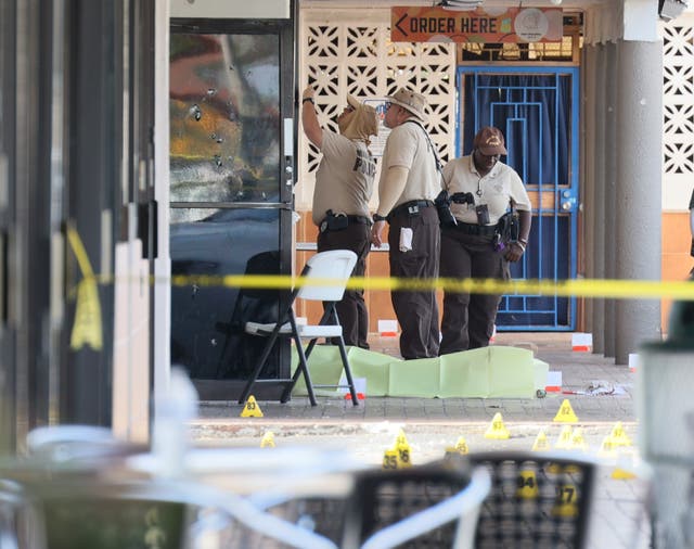 <p>Miami-Dade police investigate near shell case evidence markers on the ground and a door with what appear to be bullet holes</p>