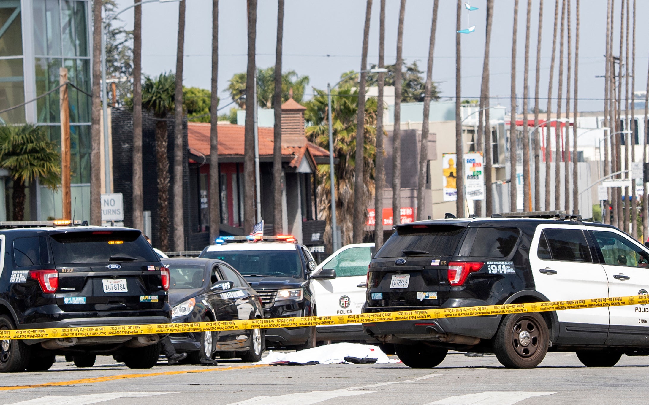 A body covered in a white sheet lies next to a black vehicle covered with stickers and a shattered driver side window, as police cars block traffic at the corner of Fairfax Avenue and Sunset Boulevard in Los Angeles on April 24, 2021 in what appears to be an officer-involved shooting.