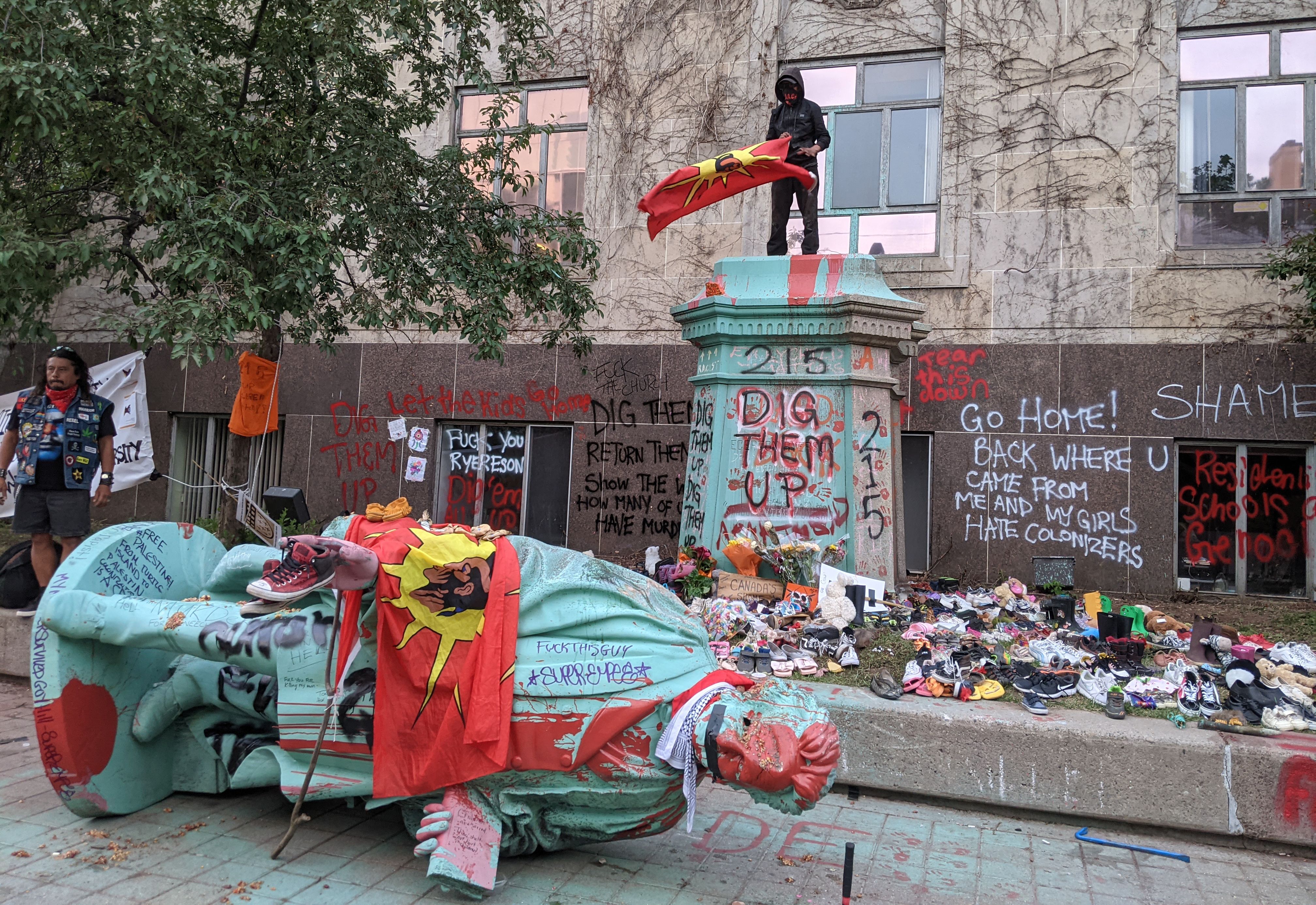 <p>A man waving the Mohawk Warrior Society flag stands on the pedestal of the toppled statue of Egerton Ryerson, one of the architects of indigenous boarding school system, in Toronto on June 6, 2021. </p>