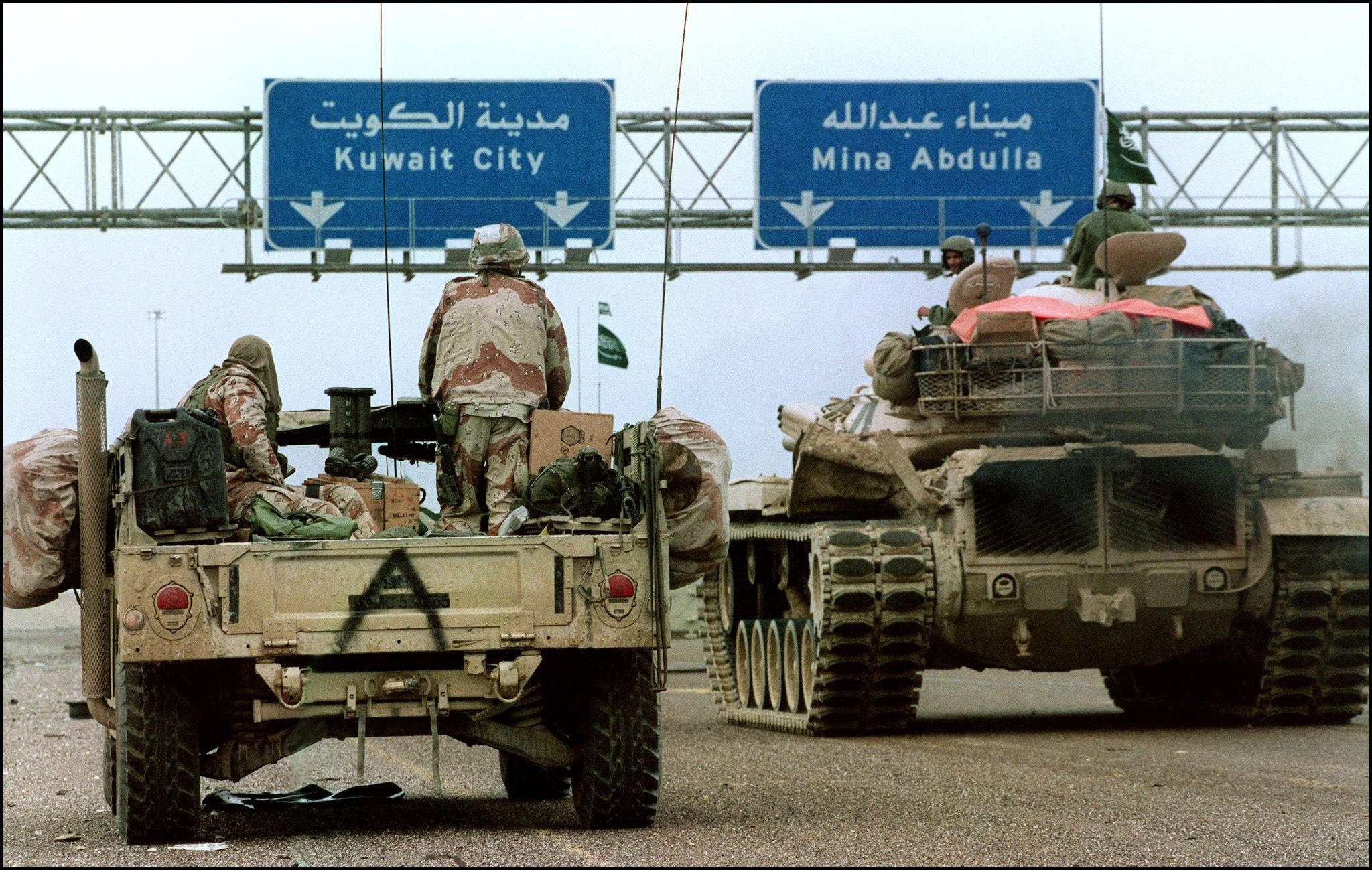 A US Humvee and a Saudi tank pass under a sign directing them to Kuwait City in February 1991 during the Desert Storm offensive