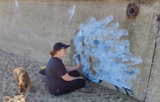 <p>A woman tries to clean the now-defaced mural of a rat in a deckchair </p>