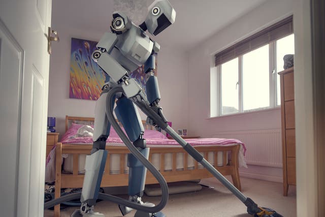 <p>A conceptual image of a generic, blue and white robot holding a vacuum cleaner doing household cleaning chores in bedroom, viewed from a low angle through an open doorway</p>