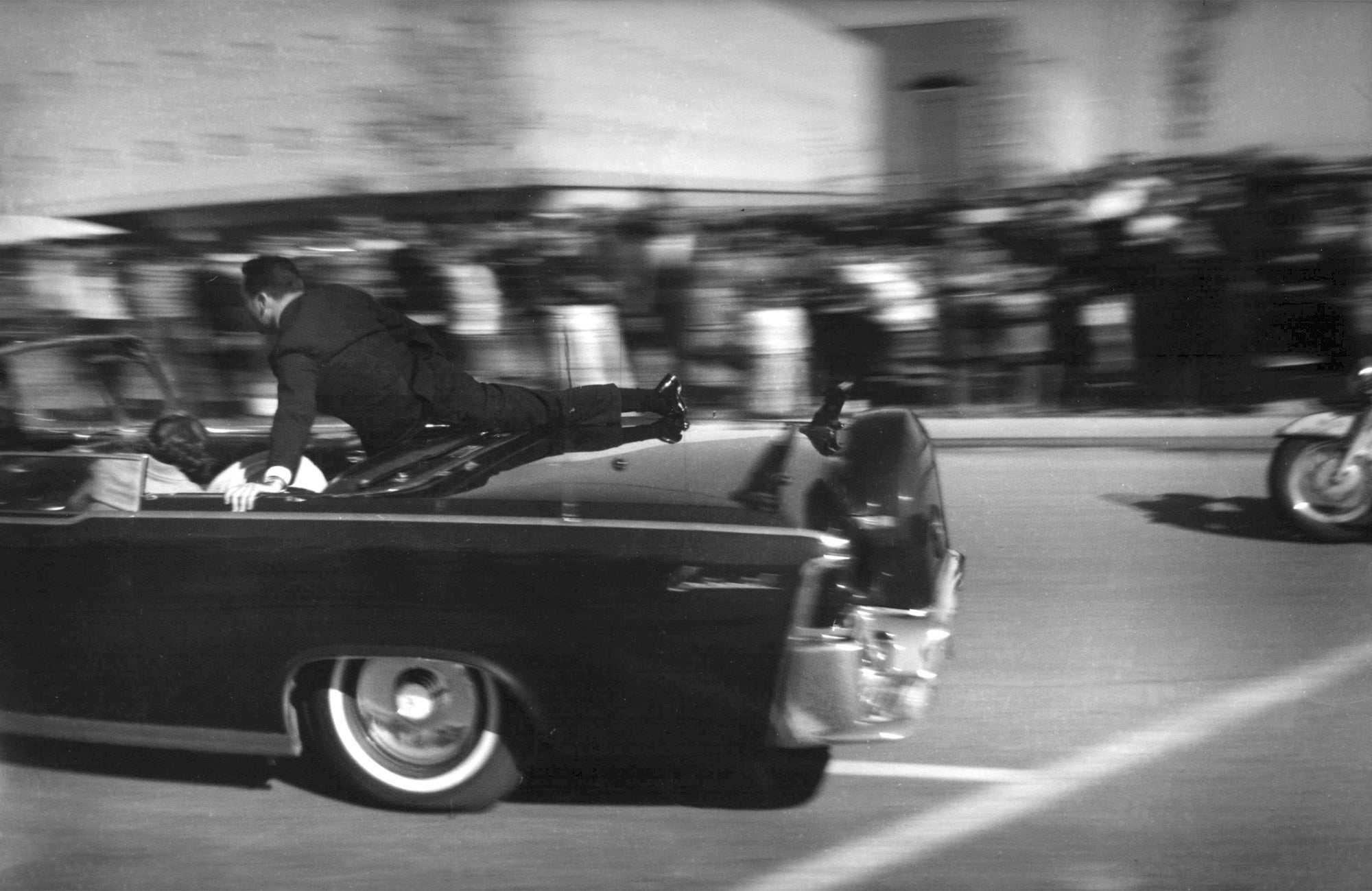 In this Nov. 22, 1963 file photo, the limousine carrying mortally wounded President John F. Kennedy races toward the hospital seconds after he was shot in Dallas. Secret Service agent Clinton Hill is riding on the back of the car, Nellie Connally, wife of Texas Gov. John Connally, bends over her wounded husband, and first lady Jacqueline Kennedy leans over the president. (AP Photo/Justin Newman, File)