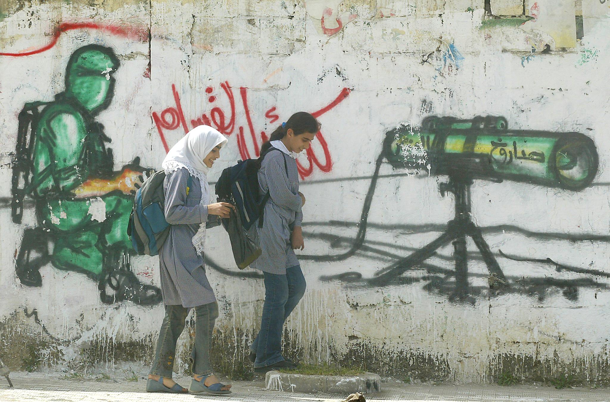 <p>Two Palestinian school girls walk past graffiti supporting Hamas. The military wing of the group has been proscribed in the UK since 2001</p>