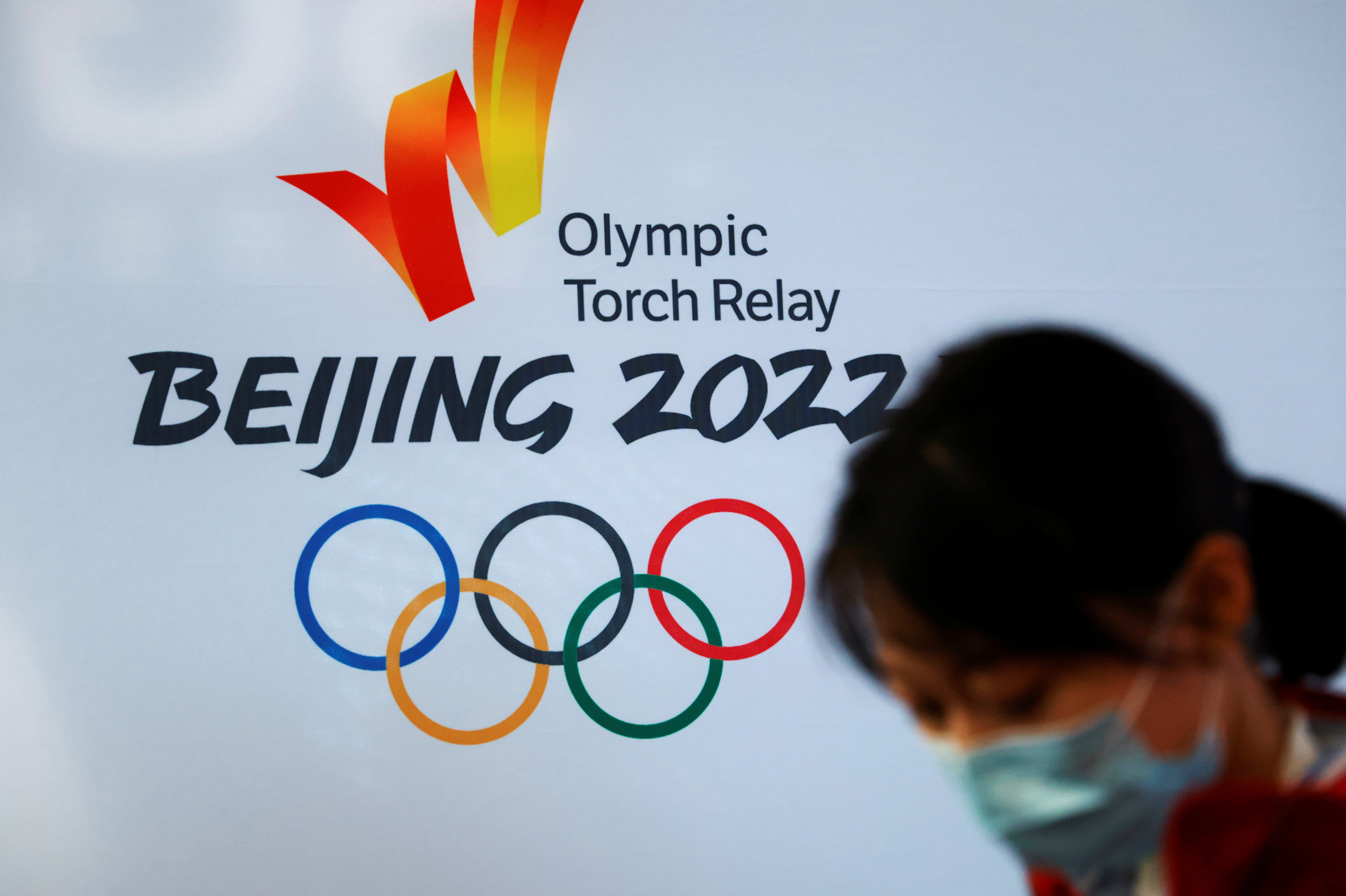 A woman wearing a face mask stands in front of the logo of the Beijing 2022 Winter Olympics before the Olympics flame exhibition tour at Beijing University of Posts and Telecommunications, in Beijing, China on 9 December 2021