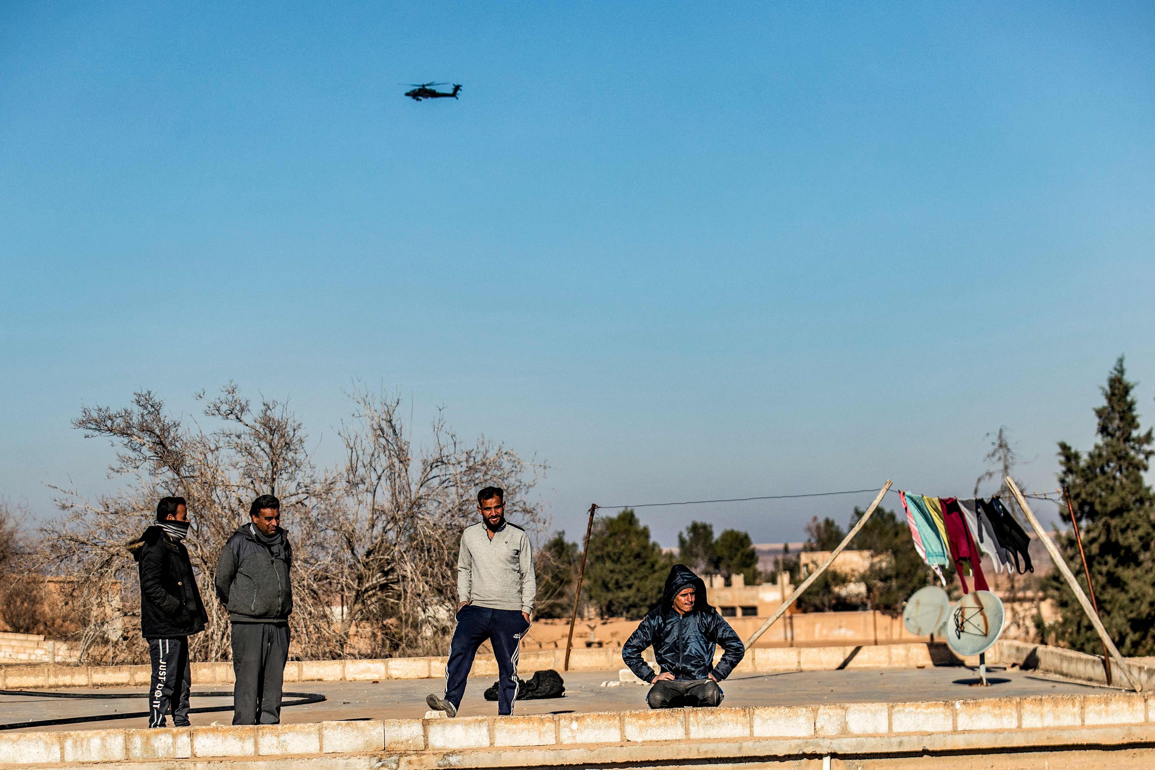 Locals watch as a A US AH-64 Apache attack helicopter flies above US soldiers patrolling along the frontlines between areas held by the Syrian Democratic Forces (SDF) and Turkish-backed fighters near the village of Dardara in the countryside of Tal Tamr