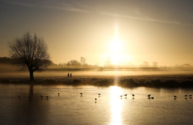 People walk through frost and mist alongside a frozen lake during sunrise in Bushy Park, London