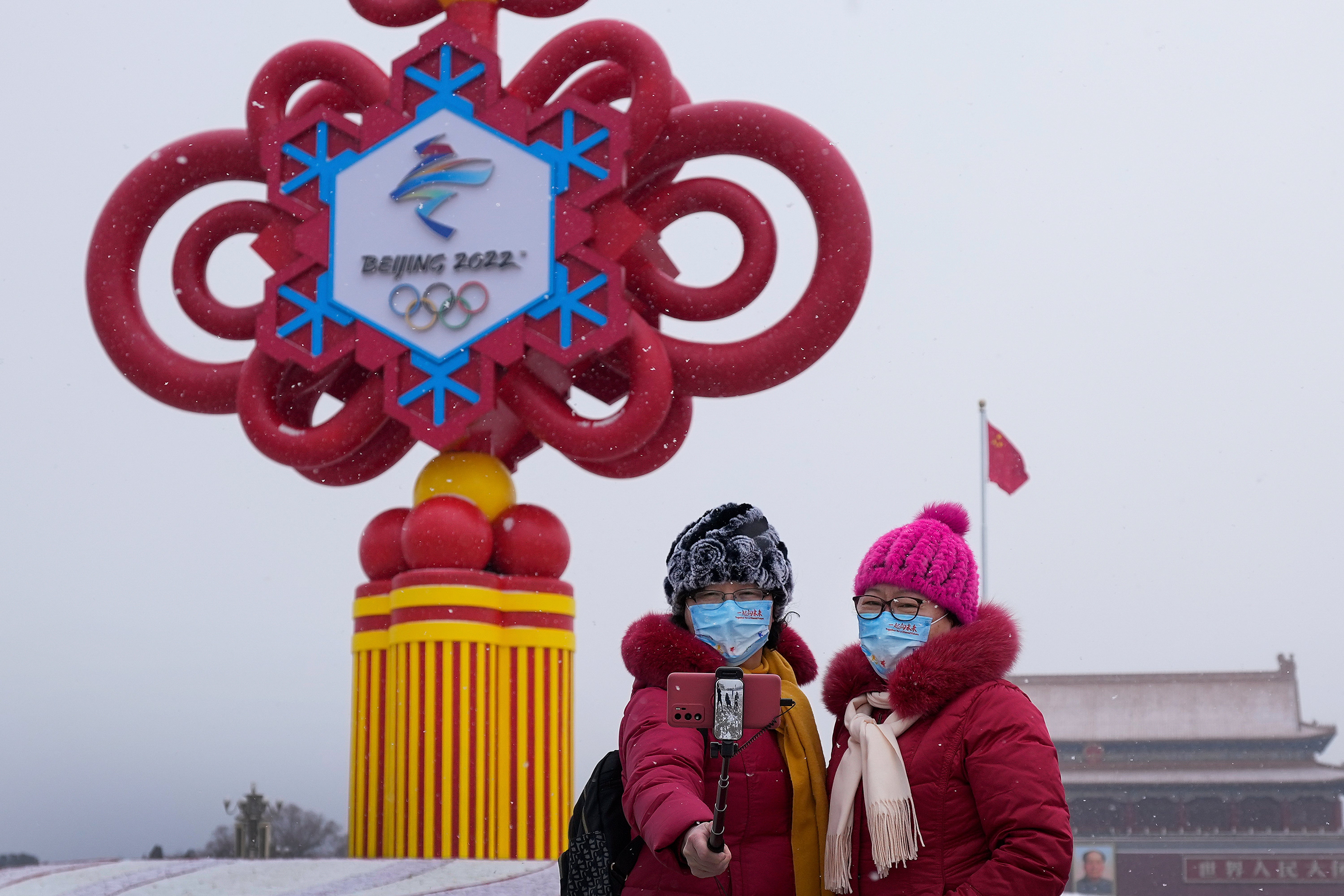 Women wearing face masks printed with a slogan for Beijing Winter Olympics Games take a selfie with a decoration for the Winter Olympics Games on Tiananmen Square in Beijing