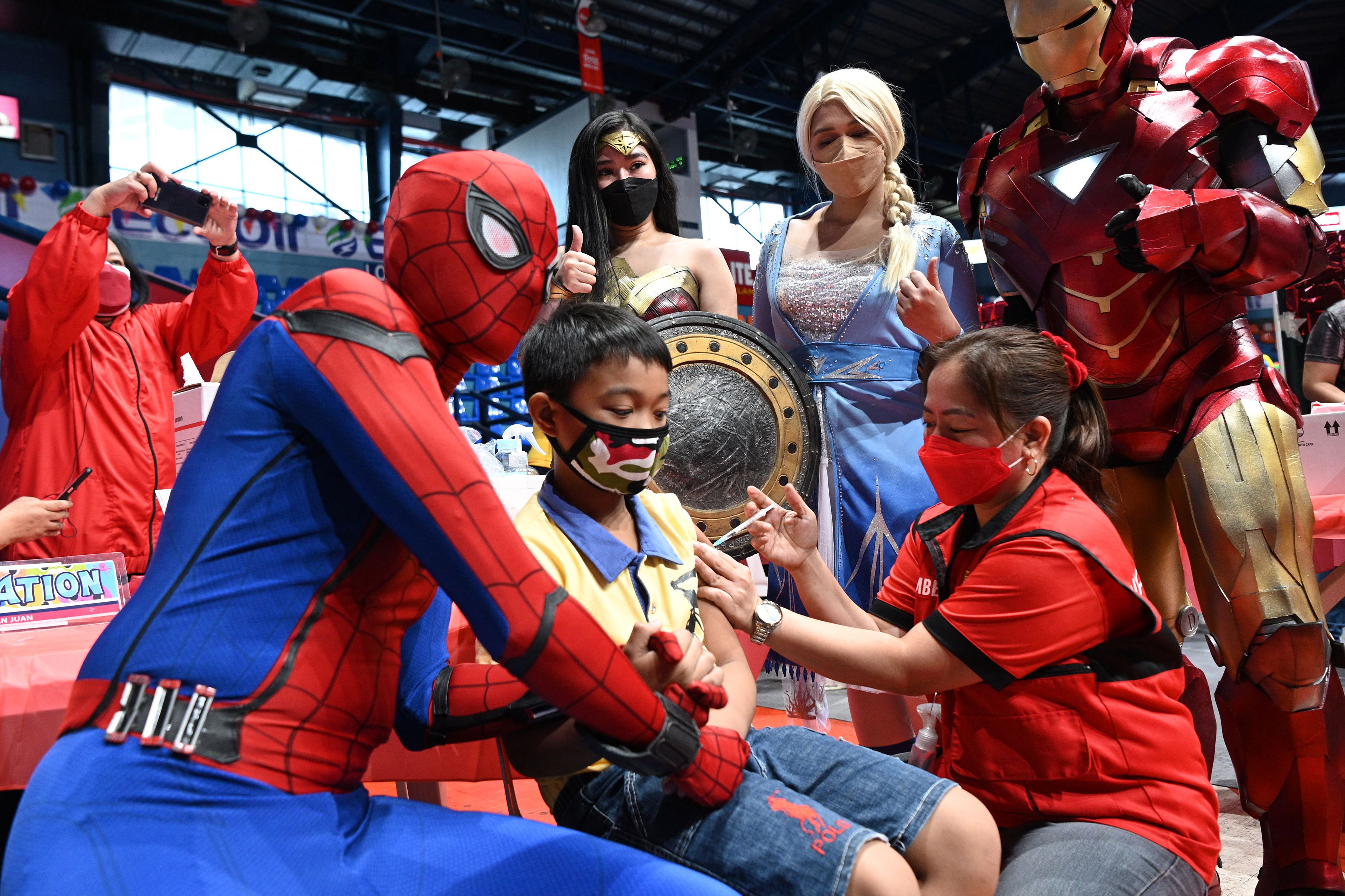 <p>Performers dressed as superhero characters pose with a child receiving the Pfizer-BioNtech Covid-19 vaccine for children aged 5-11 at a gym in San Juan City, suburban Manila</p>