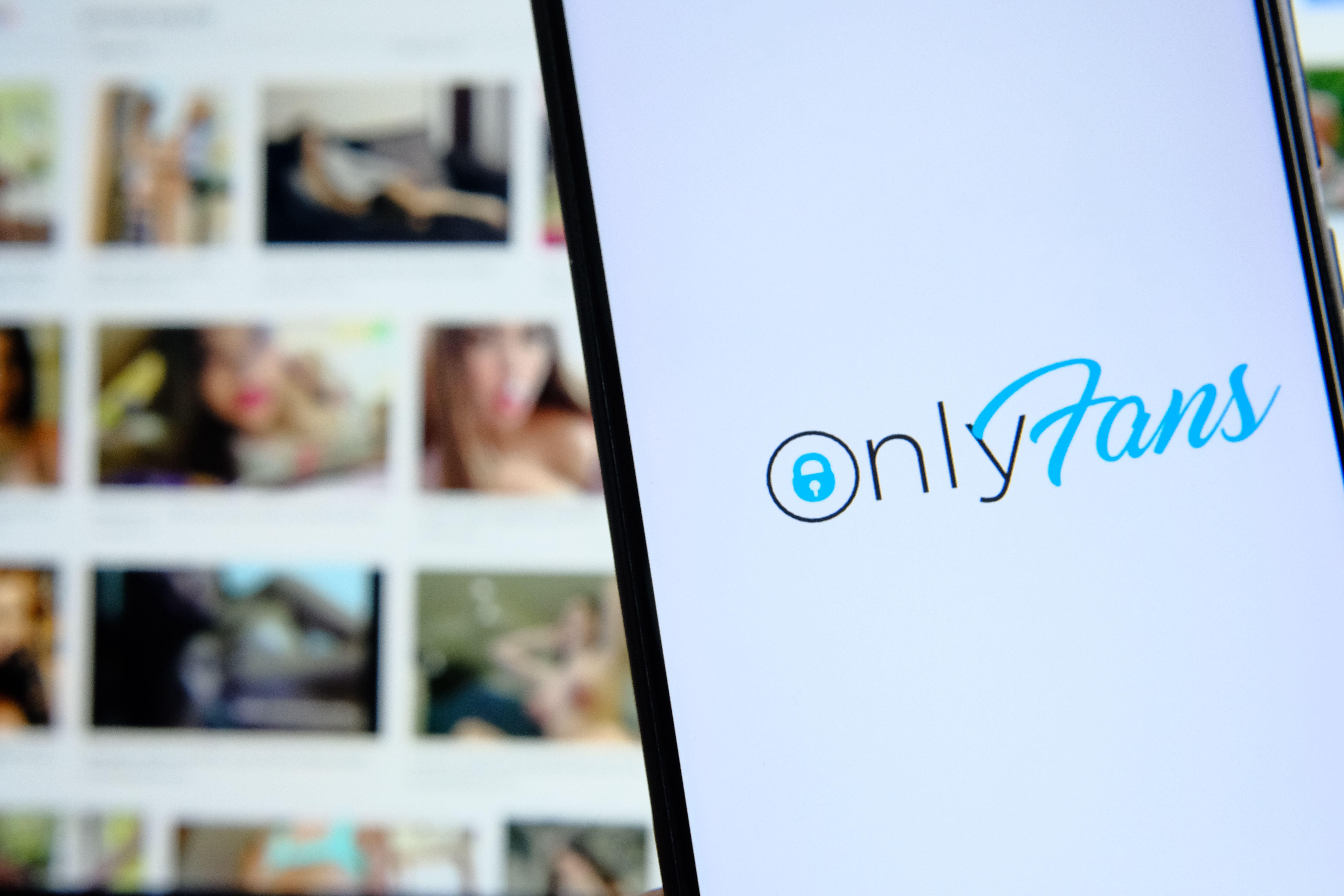 Onlyfans platform logo seen on smartphone and blurred pictures of unrecognisable girls on the background laptop