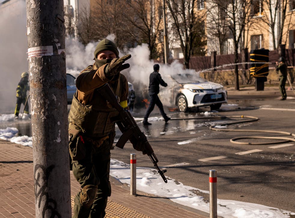 Ukrainian soldiers take positions outside a military facility as two cars burn, in a street in Kyiv, Ukraine, Saturday, Feb. 26, 2022. Russian troops stormed toward Ukraine’s capital Saturday, and street fighting broke out as city officials urged residents to take shelter. (AP Photo/Emilio Morenatti)