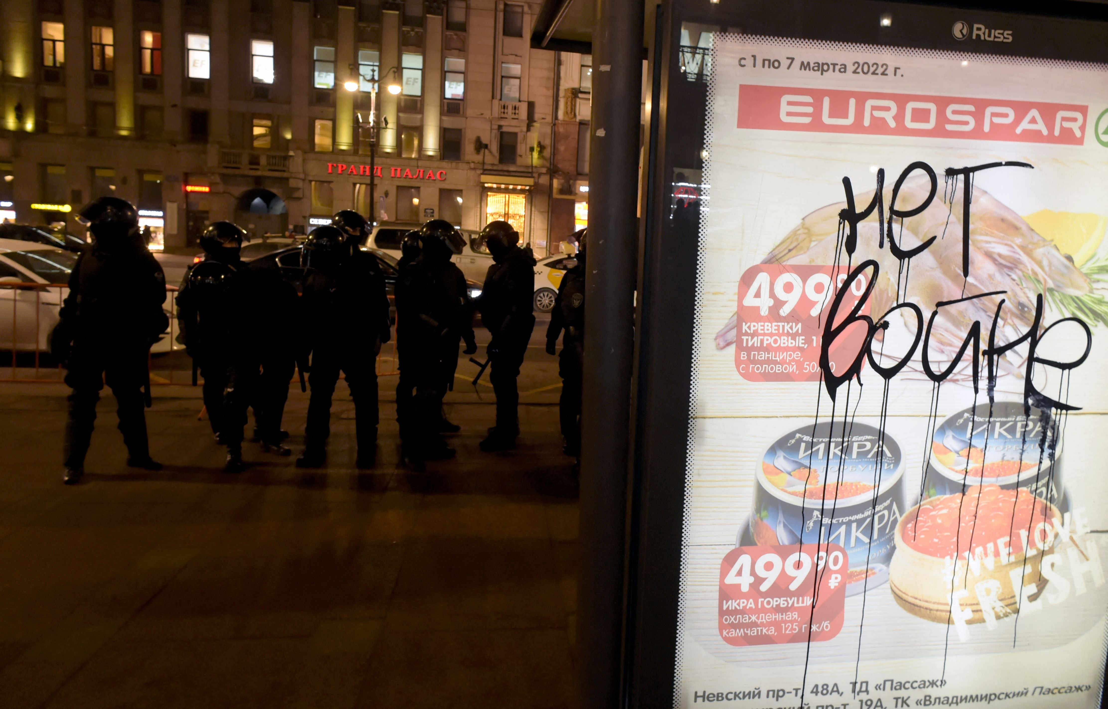 An inscription reading "No to war" is seen on an advertisement board as riot police officers stand guard nearby during a protest in Saint Petersburg.