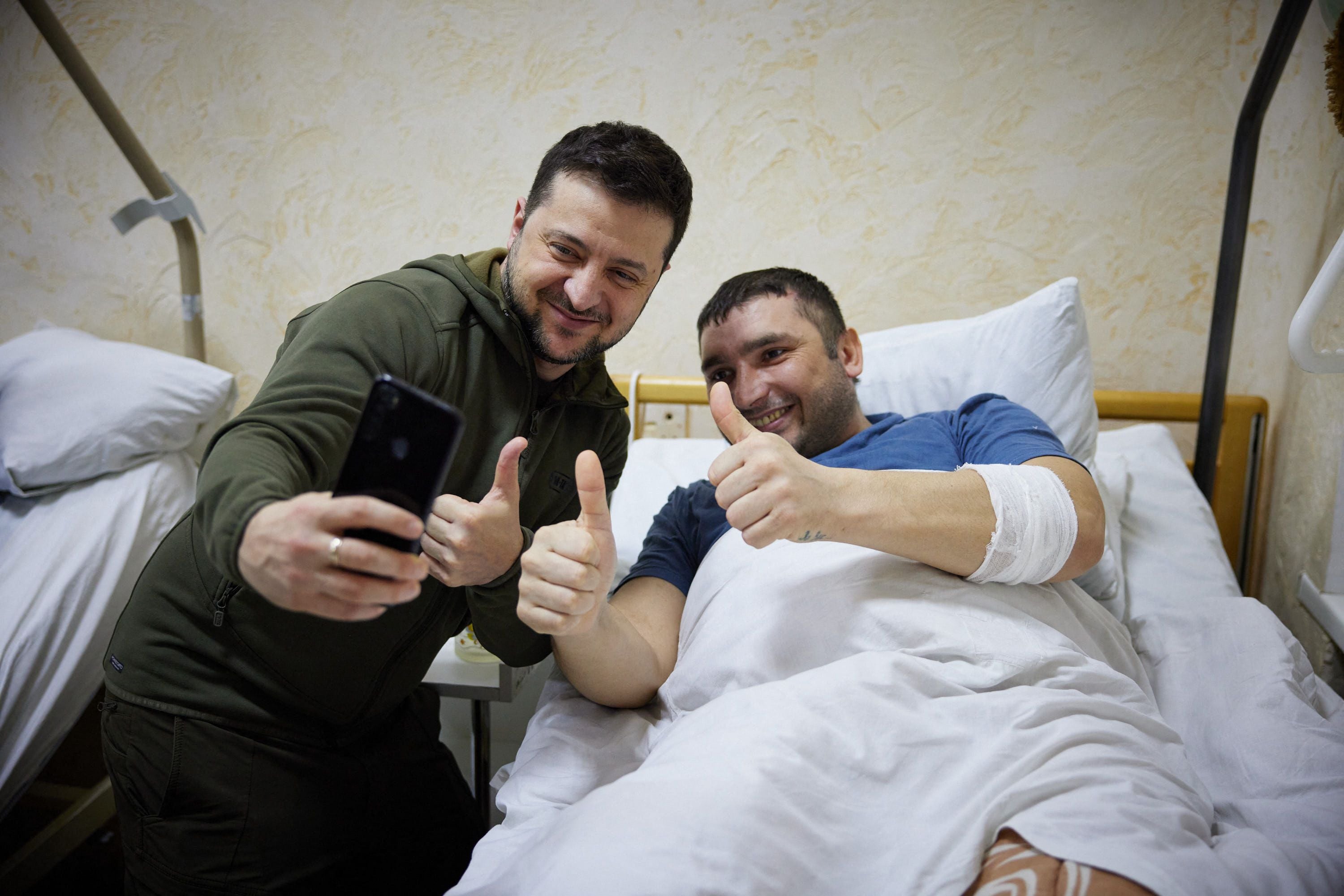 Volodymyr Zelensky snaps a selfie with an injured man laying on a bed during a visit at a military hospital following fighting in the Kyiv region