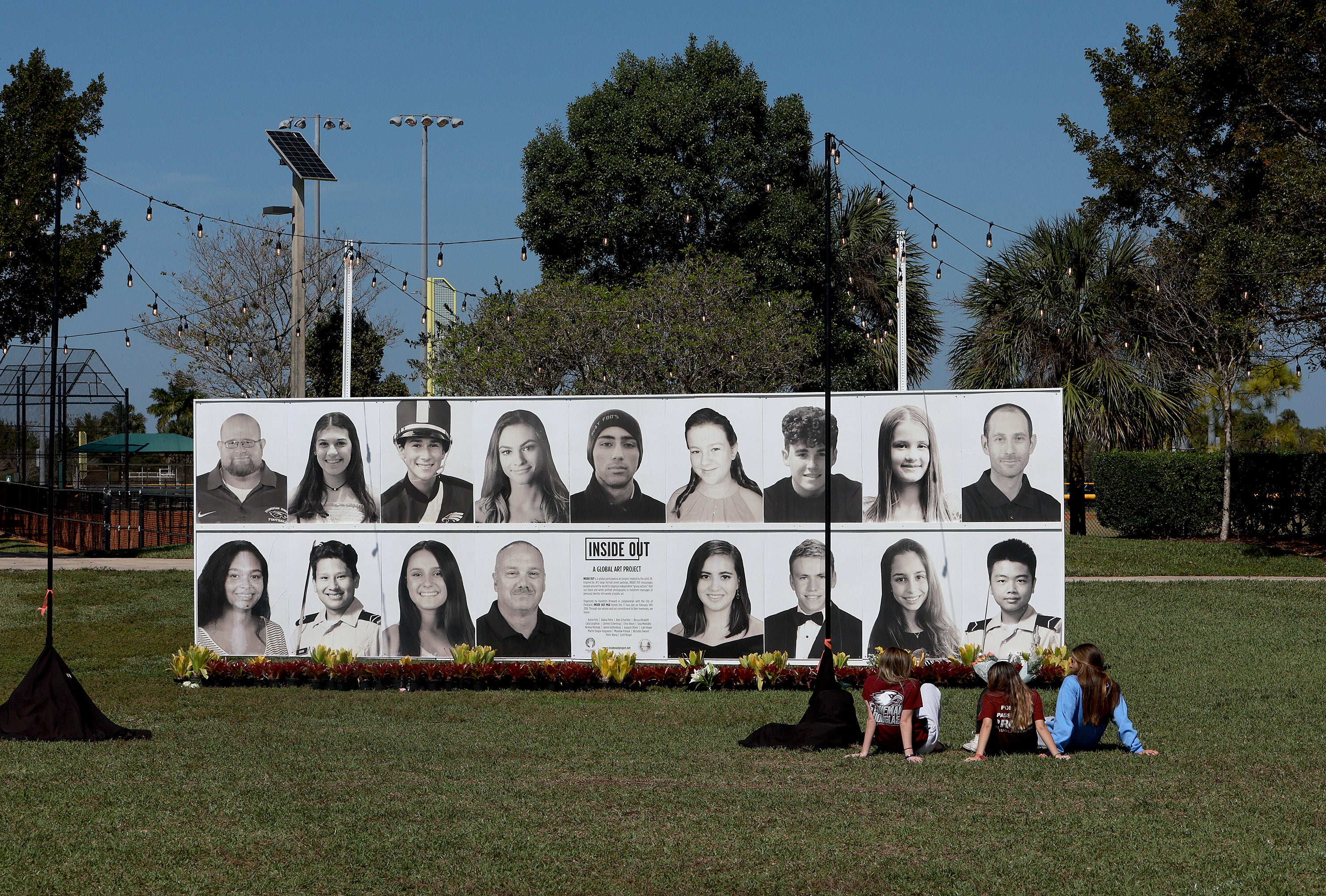 People sit in front of a photo display of the 17 people killed at Marjory Stoneman Douglas High School during a mass shooting on February 14, 2022 in Pine Trails Park in Parkland, Florida