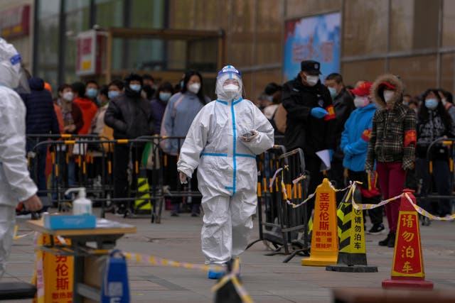 <p>A health worker wearing a protective suit walks by masked residents who wait in line to get their throat swab at a coronavirus testing site in Beijing on 6 April</p>