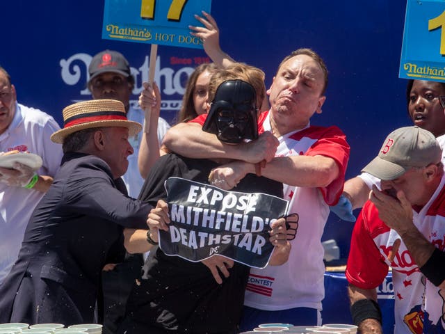 <p>Top competitive eater Joey Chestnut (C) tackles a protester with a ‘Expose Mithfield’s Deathstar’ sign during Nathan’s Famous Fourth of July International Hot Dog Eating Contest at Coney Island in New York, New York, USA, 4 July 2022</p>