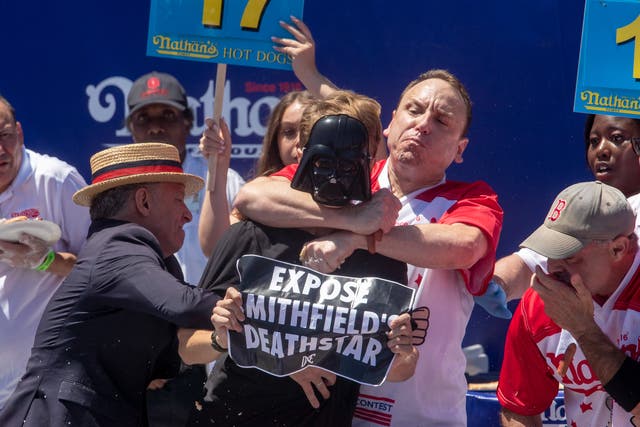 <p>Top competitive eater Joey Chestnut (C) tackles a protester with a ‘Expose Mithfield’s Deathstar’ sign during Nathan’s Famous Fourth of July International Hot Dog Eating Contest at Coney Island in New York, New York, USA, 4 July 2022</p>
