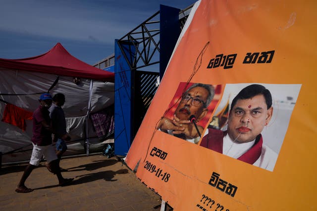 <p>People walk past a poster showing defaced portraits of president Gotabaya Rajapaksa and his brother Basil at the ongoing protest site outside the president’s office</p>