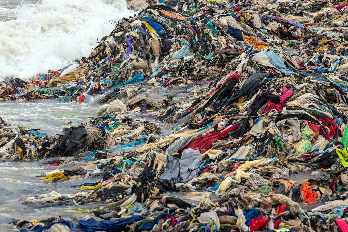Massive piles of discarded textiles on a shoreline, illustrating wealthy cities exporting waste to the Global South and its environmental impact