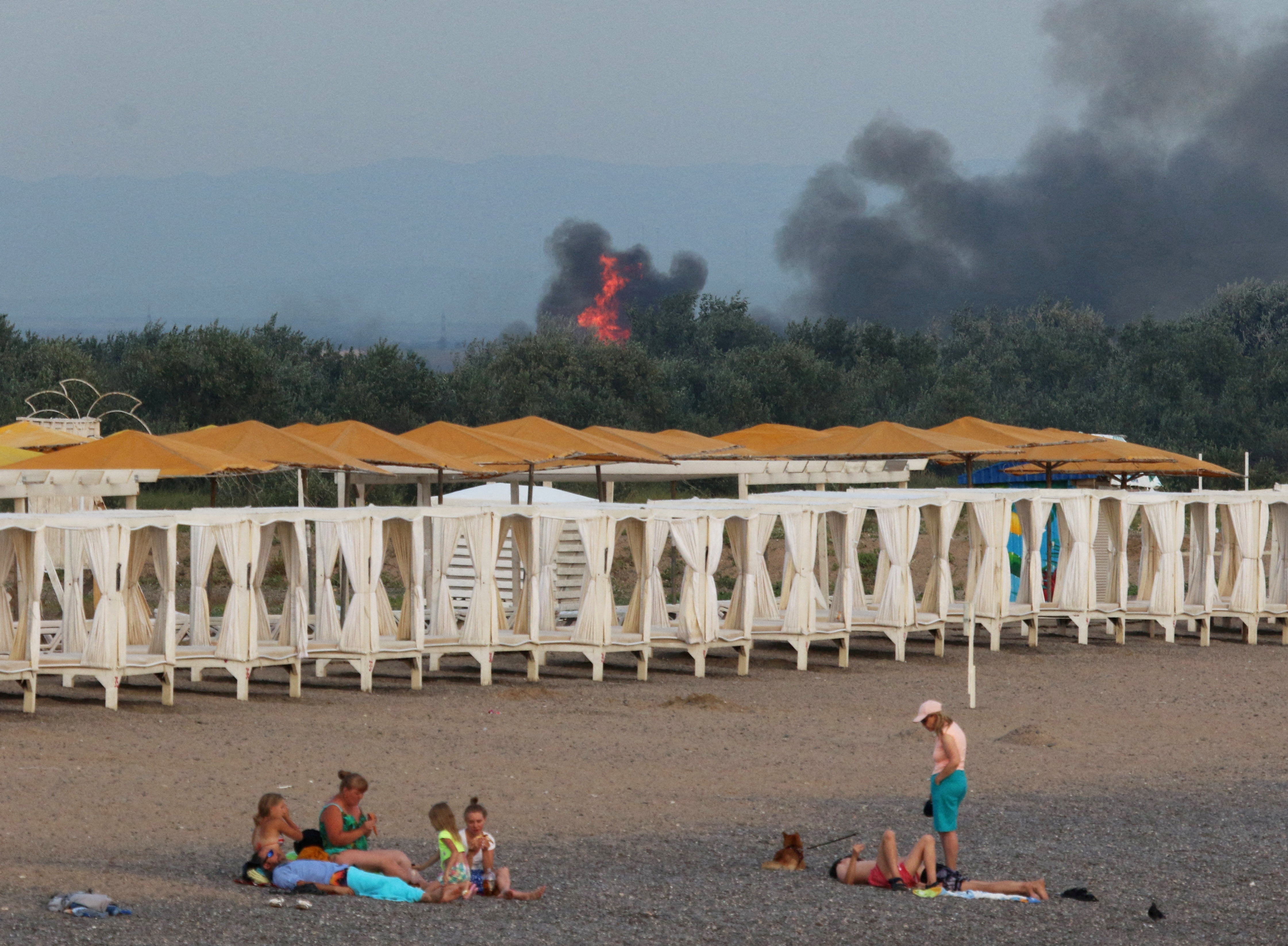 People rest on a beach in Novofedorivka as smoke rises following explosions at a Russian military airbase