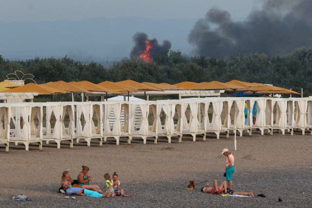 <p>People rest on a beach as smoke and flames rise after explosions at a Russian military airbase, in Novofedorivka</p>