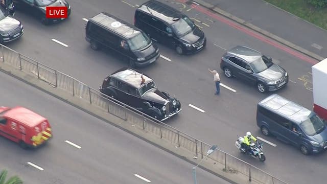 Screengrab from BBC News of a member of the public stepping out into the road towards a car carrying King Charles III and the Queen Consort to take a photo with close protection officers leaving a car which is providing security and gesturing to the member of public (BBC/PA)