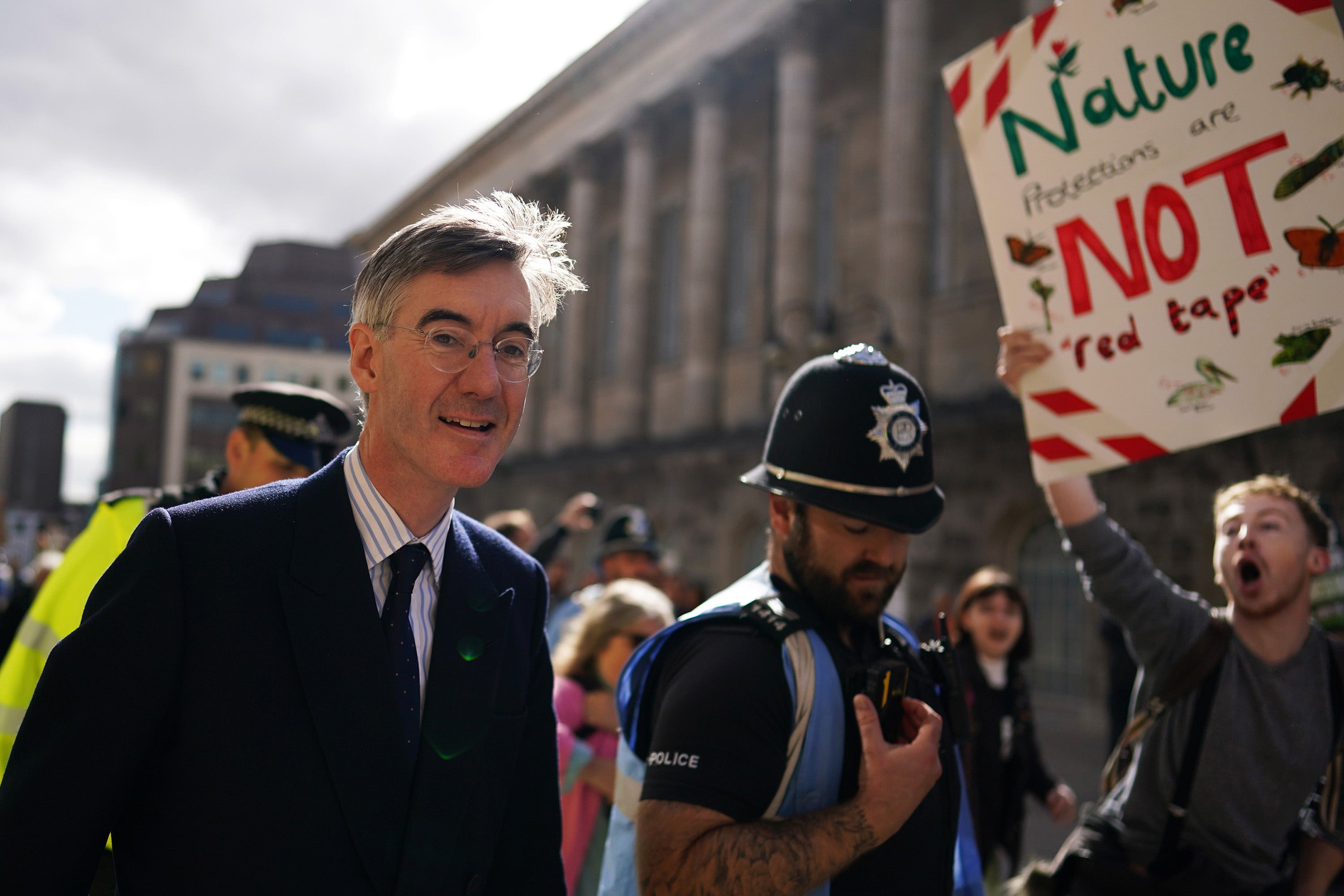 A smiling Jacob Rees-Mogg surrounded by police, walks through protesters in Birmingham