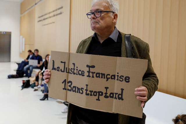 <p>Wilfried Schmid, a family member of a victim, holds a placard that reads ‘French Justice 13 years too late’ </p>