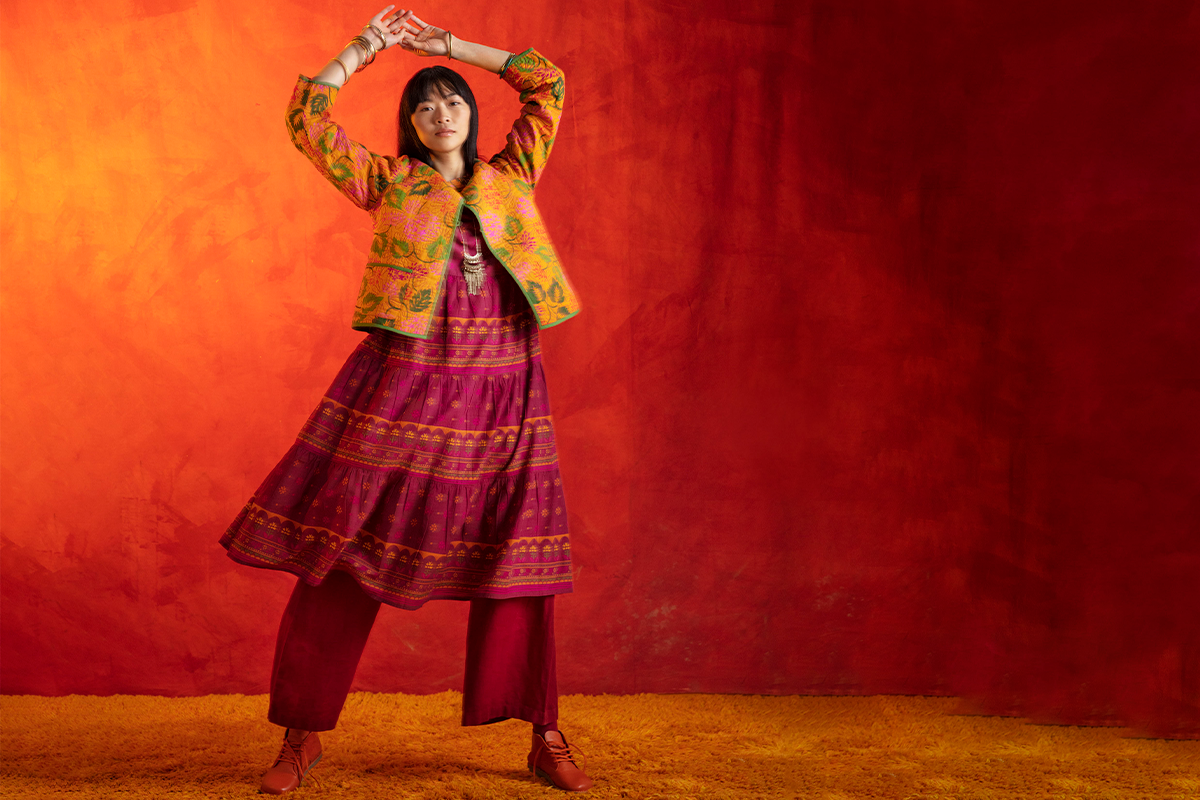 Woman wearing colourful textiles with her arms raised against a red backdrop