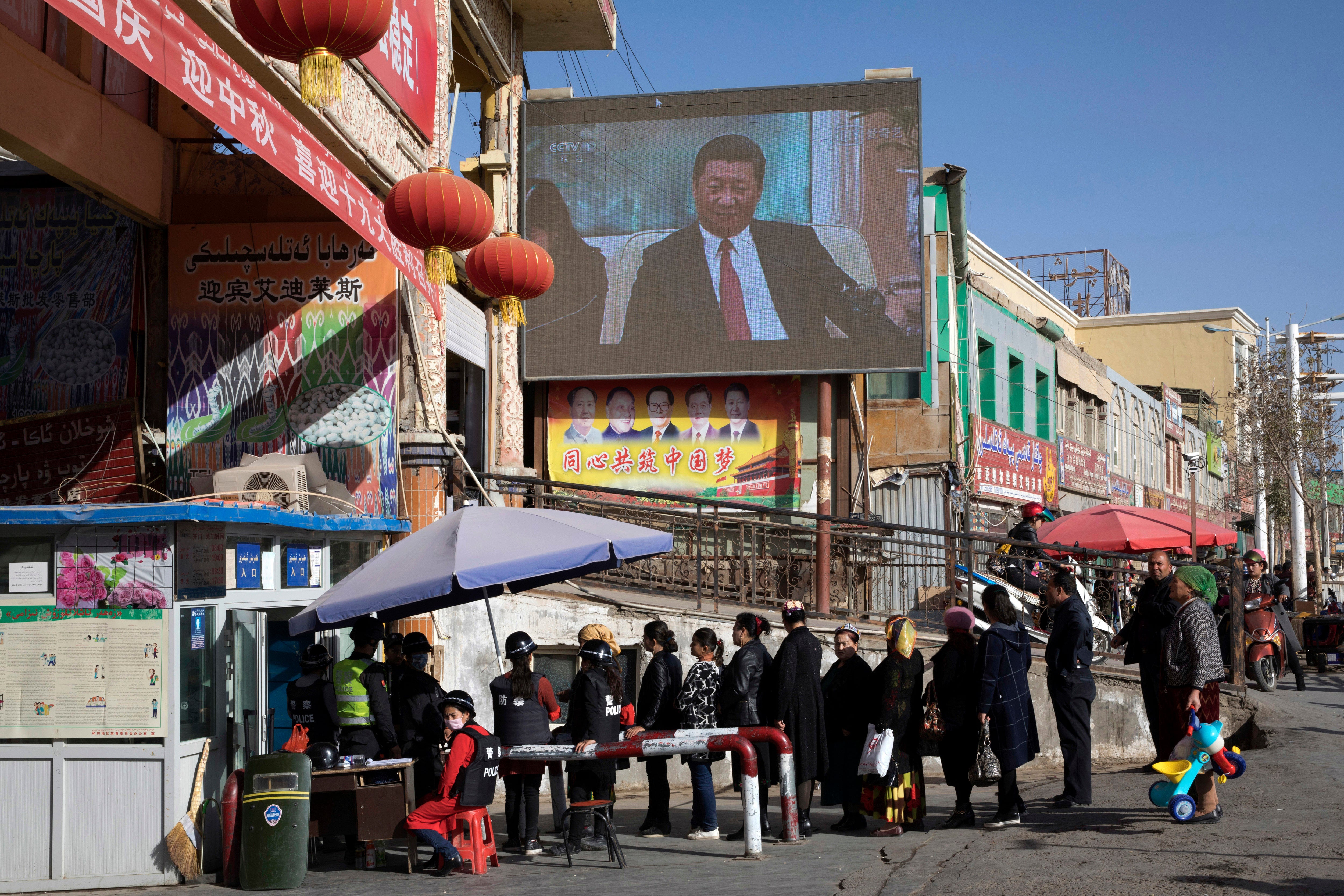 Residents walk through a security checkpoint into a bazaar where a screen shows China’s president Xi Jinping in Hotan in Xinjiang