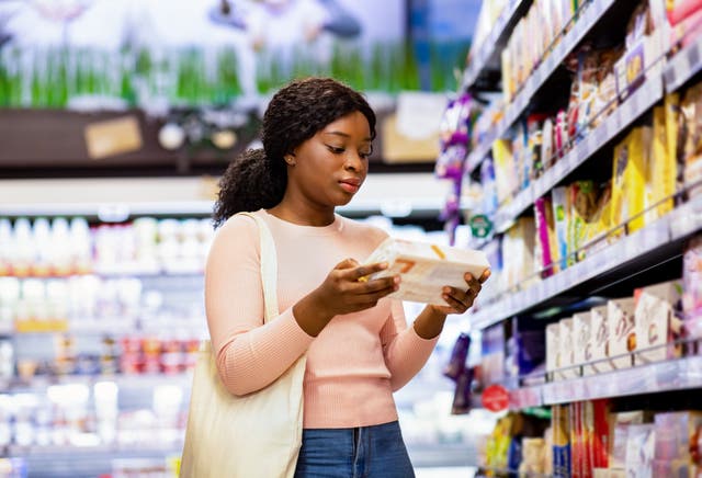 <p>Attractive black woman with tote bag holding food product, buying groceries at supermarket. Beautiful African American lady looking through labels at grocery department of huge mall</p>