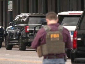 An FBI agent stands in front of a standoff between police and a man in a car with a gun to his head