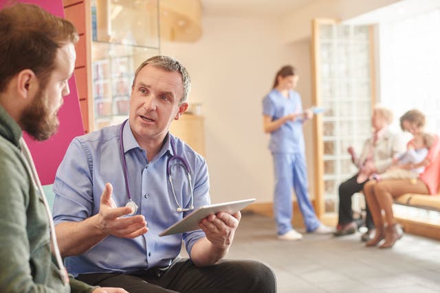 <p>A mature male doctor sits next to a male patient in his waiting room and casually chats through a few things on his digital tablet . In the background a nurse chats to a mother and child and grandmother with her digital tablet .</p>